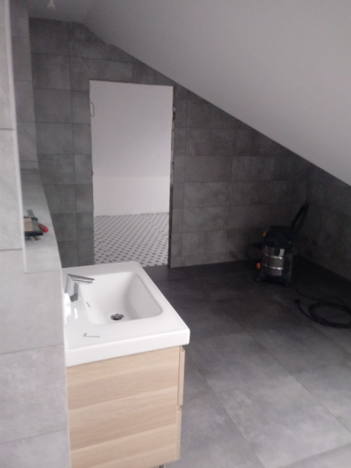 Umywalka z szafką w szarej łazience na poddaszu, widoczny odkurzacz i płytki w innym pomieszczeniu