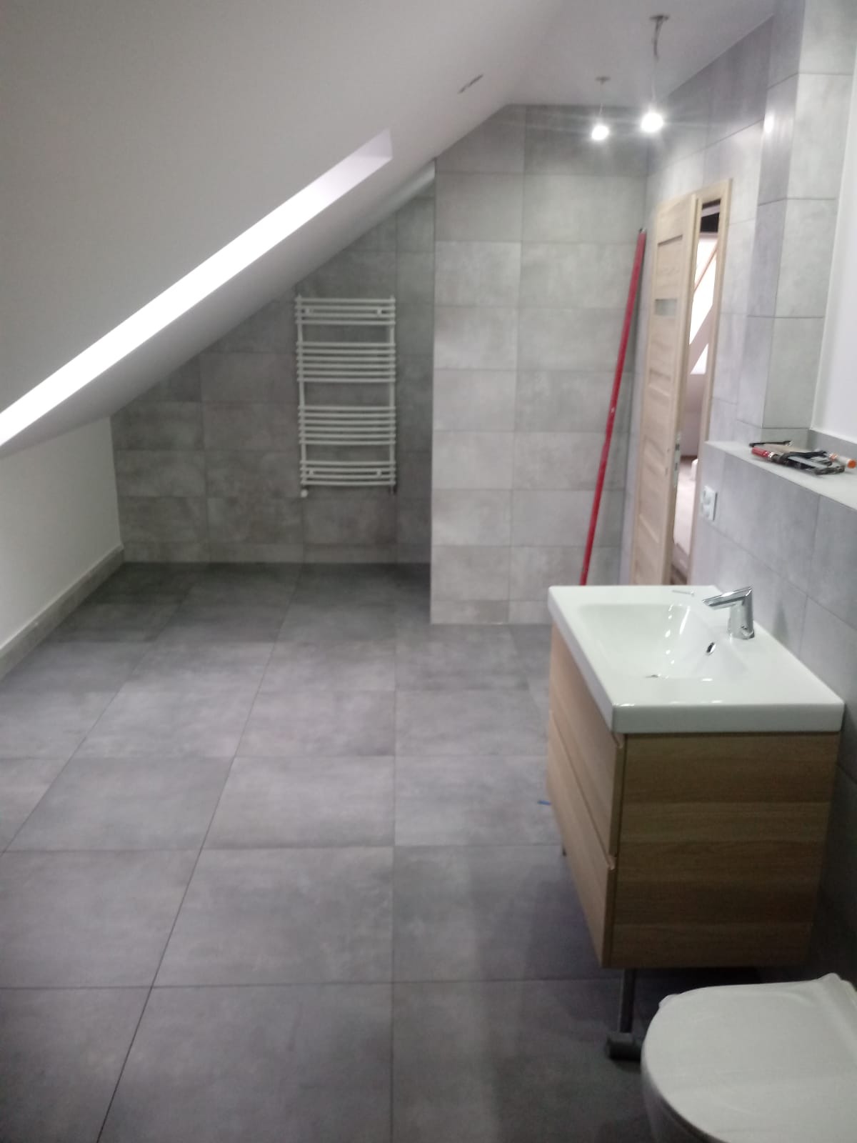 Wykończona łazienka na poddaszu z szarymi płytkami, drewnianą szafką pod umywalką i grzejnikiem drabinkowym, widoczna poziomica oparta o ścianę.