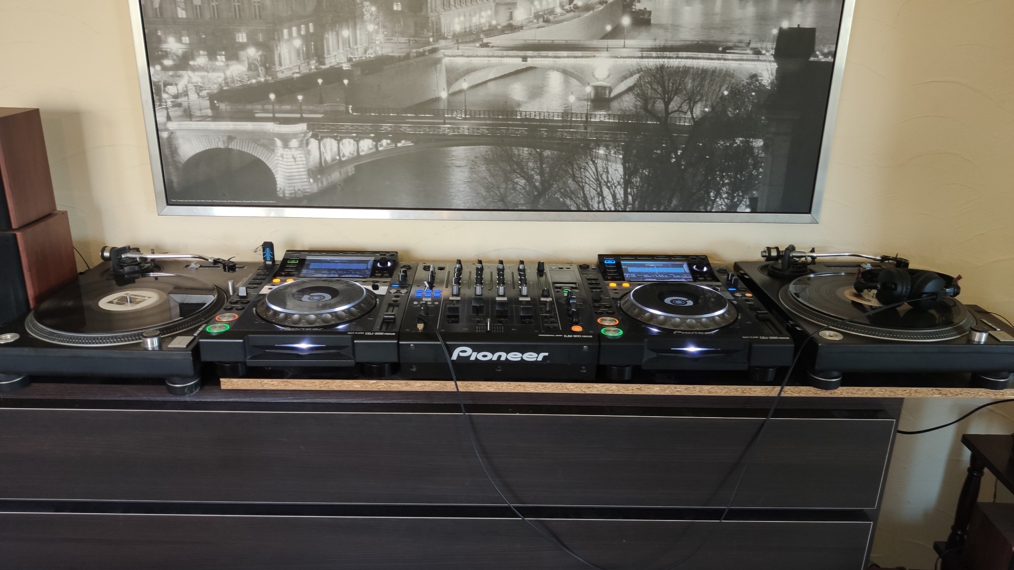 Profesjonalny sprzęt DJski Pioneer: dwa gramofony, dwa odtwarzacze CDJ-2000 Nexus i mikser DJM-900 Nexus, ustawione na komodzie, nad nimi czarno-białe zdjęcie mostu w Paryżu.