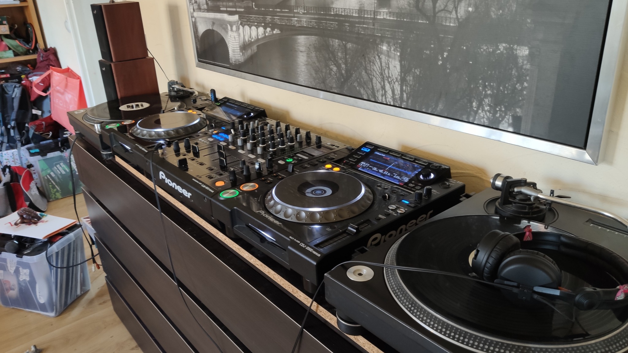 Profesjonalny sprzęt DJ-ski marki Pioneer, składający się z dwóch gramofonów i miksera, ustawiony na ciemnej komodzie w pomieszczeniu z obrazem mostu w tle.