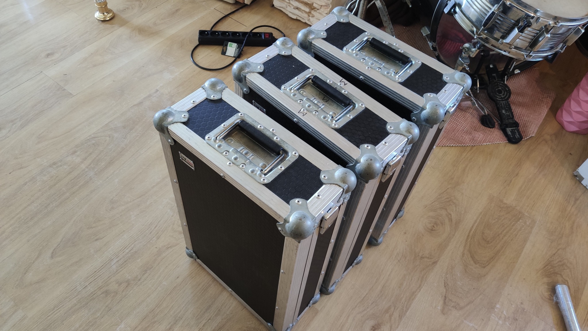 Trzy skrzynie transportowe z metalowymi okuciami, ustawione na drewnianej podłodze, obok perkusji i listwy zasilającej.