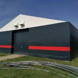 Zewnętrzny widok hali namiotowej z szarej plandeki z czerwonym pasem, biały dach z dwoma wentylatorami, rury na trawie sugerujące montaż.