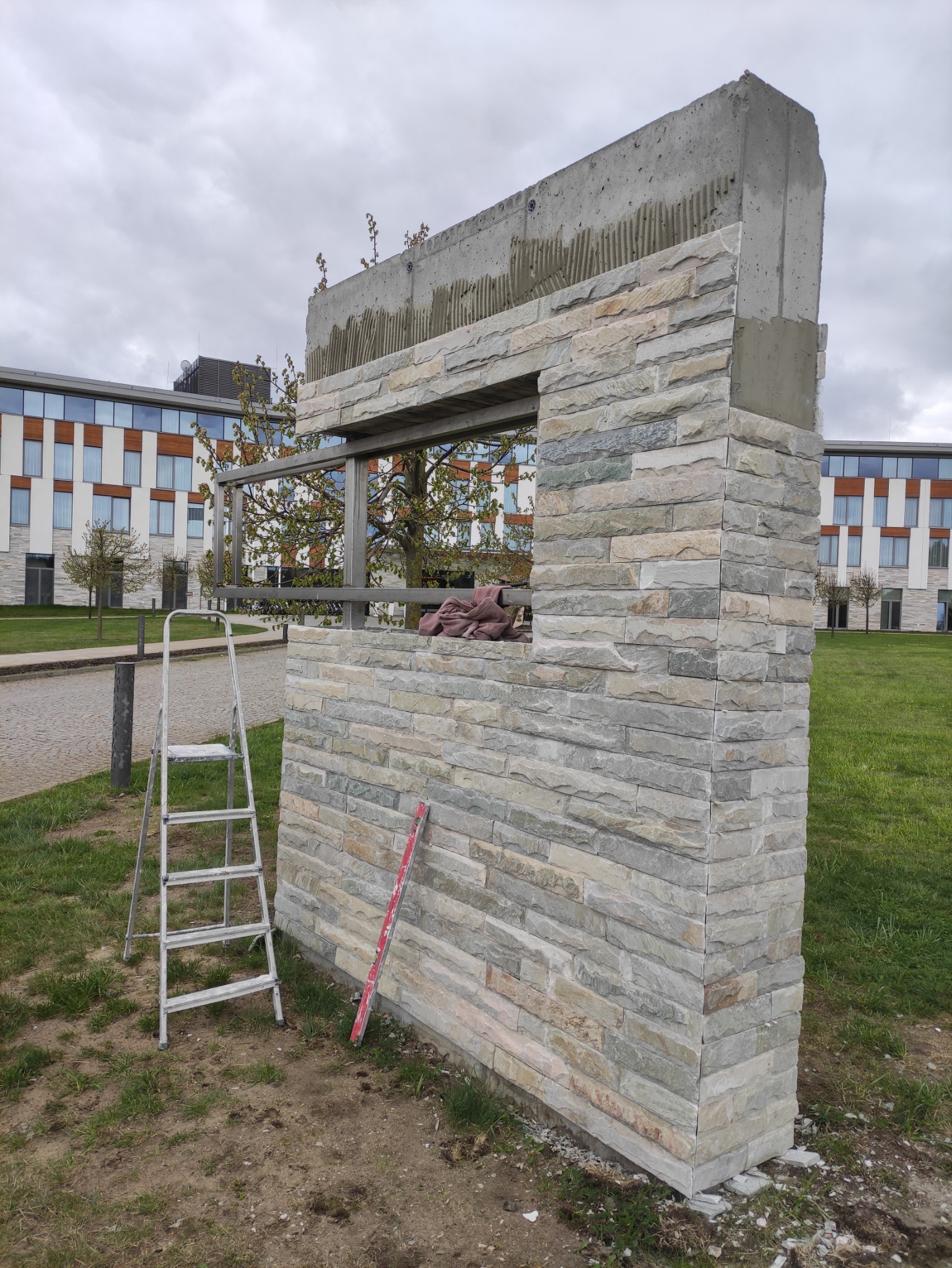 Fragment ściany z kamienia elewacyjnego w trakcie montażu, widoczny fragment surowego betonu z nałożonym klejem, drabina oparta o ścianę, poziomnica w kolorze czerwonym.