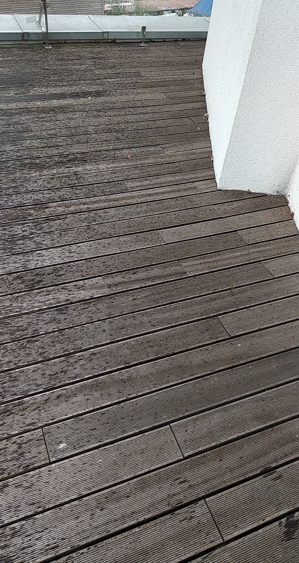 Drewniana podłoga tarasu z widocznymi śladami wilgoci, ułożona równolegle do białej ściany w tle.