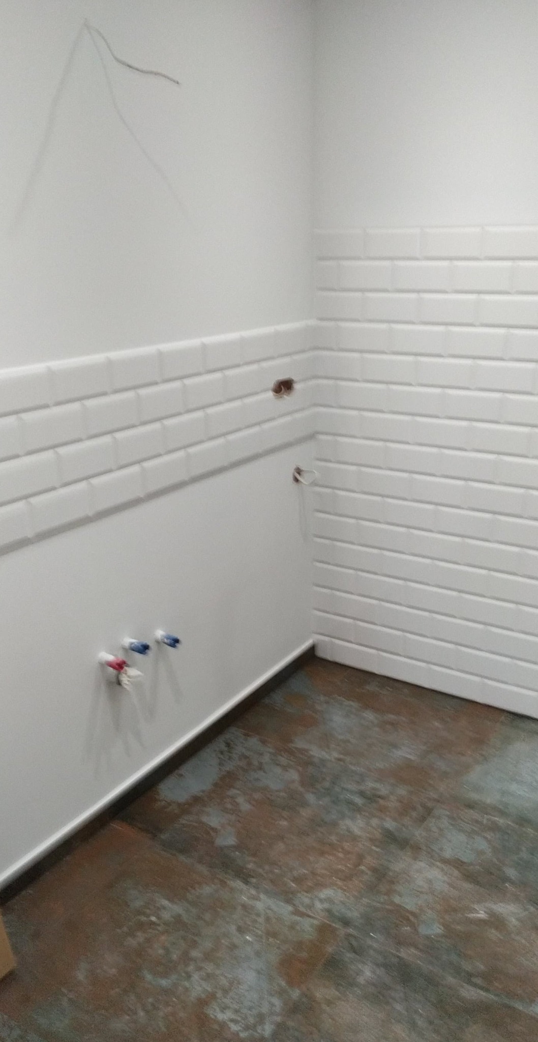 Ściana w trakcie remontu kuchni z częściowo ułożonymi białymi płytkami typu metro, wystającymi przewodami i rurami oraz surową, niejednolitą podłogą.