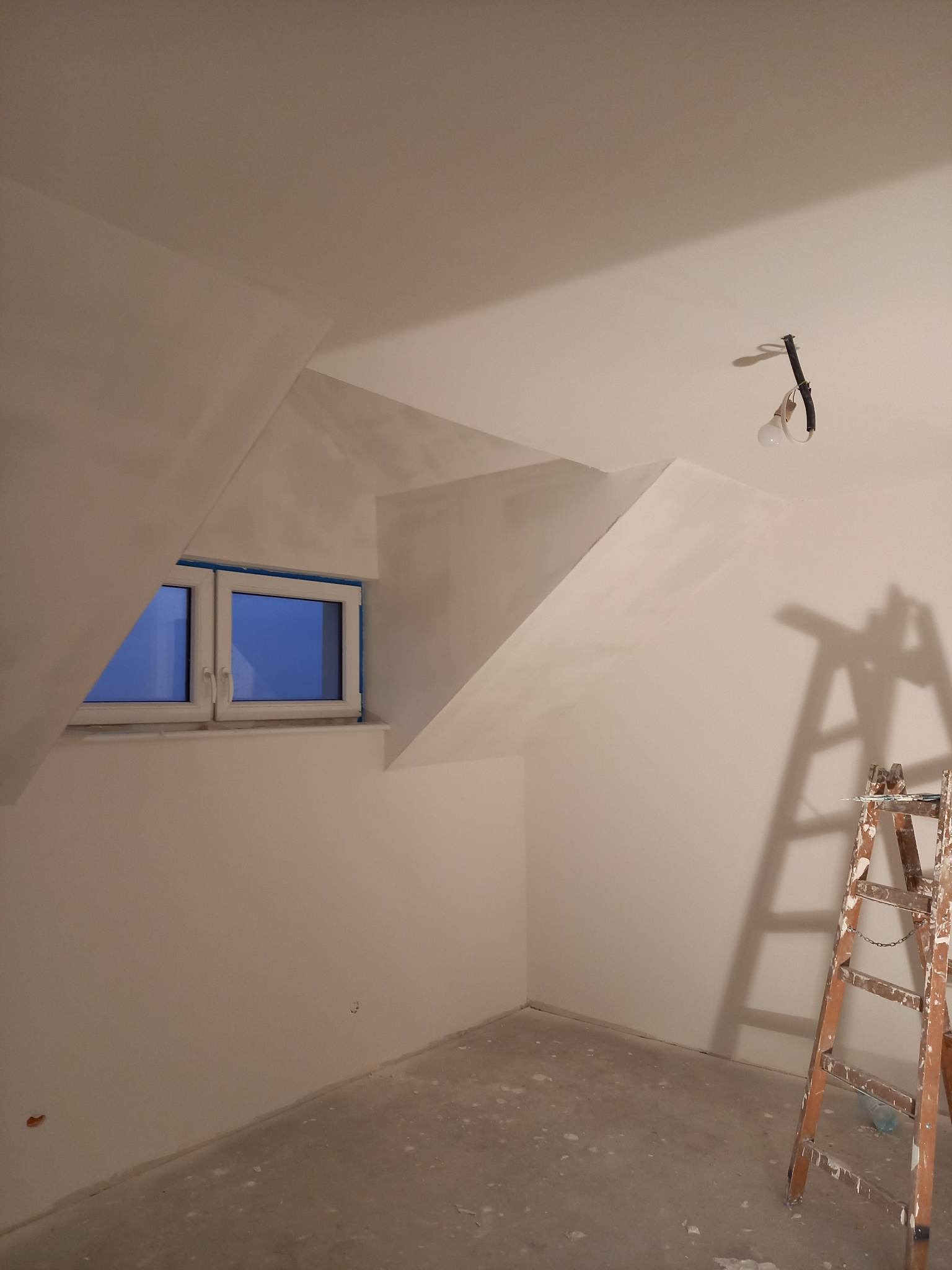 Pokój na poddaszu w trakcie remontu, ściany świeżo wygładzone, okno zabezpieczone folią, drabina malarska w rogu pomieszczenia, widoczne ślady gładzi na podłodze.