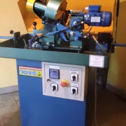 Stacjonarna szlifierka do ostrzenia narzędzi stolarskich z żółtą osłoną i niebieską podstawą, widoczne elementy sterujące i ostrzegawcze naklejki.