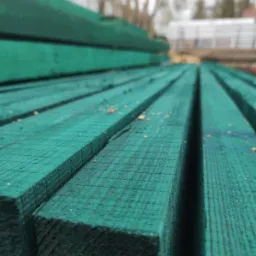 Stos impregnowanych na zielono kantówek z widoczną fakturą drewna i drobnymi wiórami, ułożonych równolegle w plenerze, z rozmytym tłem drzew i budynku.