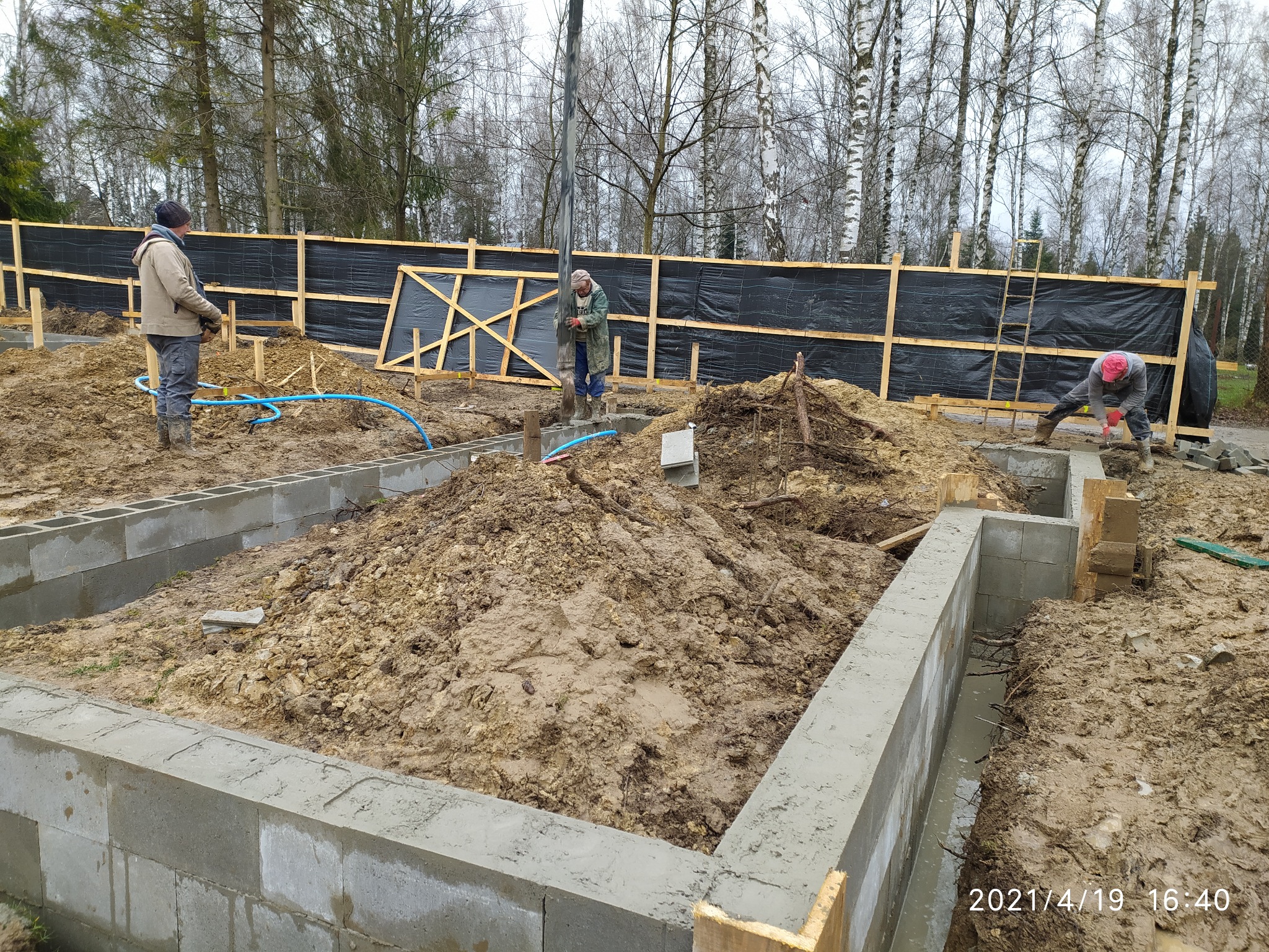 Wykopane fundamenty pod budowę domu, z widocznymi bloczkami fundamentowymi, w tle trzech pracowników i drewniane szalunki z czarną folią.