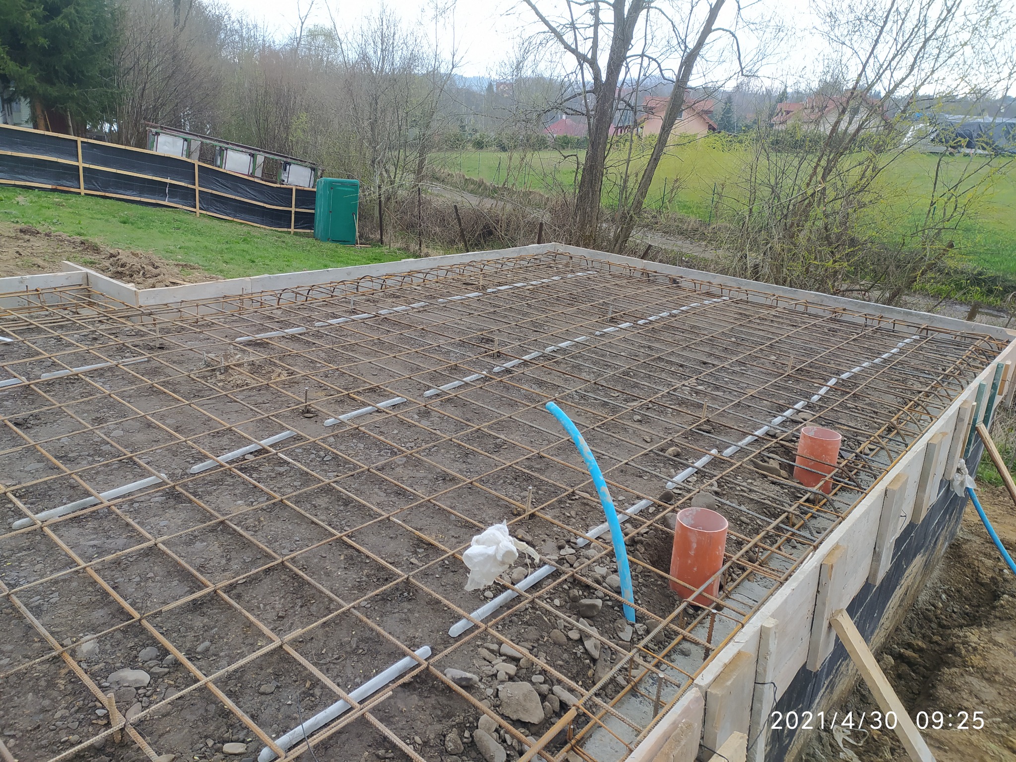 Przygotowanie zbrojenia płyty fundamentowej z widocznymi rurami instalacyjnymi, szalunkiem i elementami izolacji pionowej, w tle zielony teren z drzewami.