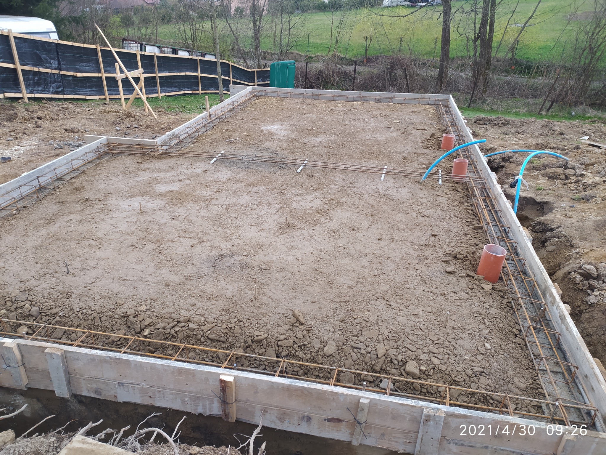 Przygotowany teren pod fundament domu z drewnianym szalunkiem, zbrojeniem i wystającymi rurami instalacji wodnej, widoczne zabezpieczenie terenu budowy w tle.