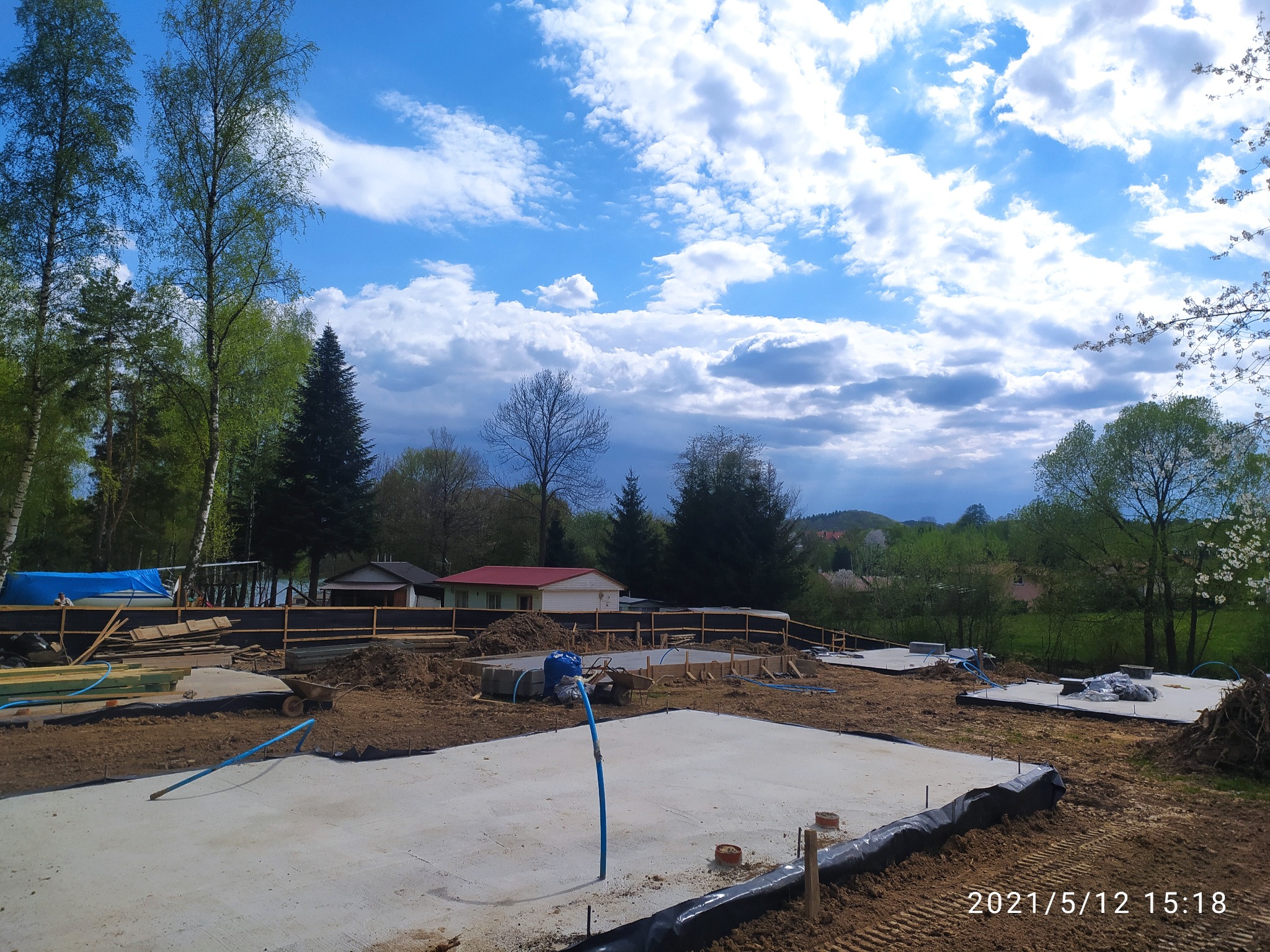 Wylana płyta fundamentowa z wystającymi rurami instalacyjnymi, otoczona ziemią i deskami szalunkowymi, w tle zabudowa jednorodzinna oraz drzewa pod błękitnym niebem z chmurami.