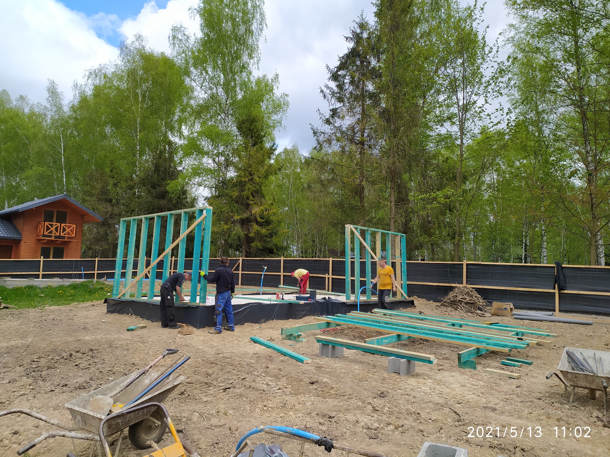 Montaż szkieletu drewnianego domu na fundamencie, widoczni pracownicy w trakcie pracy, przygotowane elementy konstrukcyjne i narzędzia na placu budowy w otoczeniu drzew.