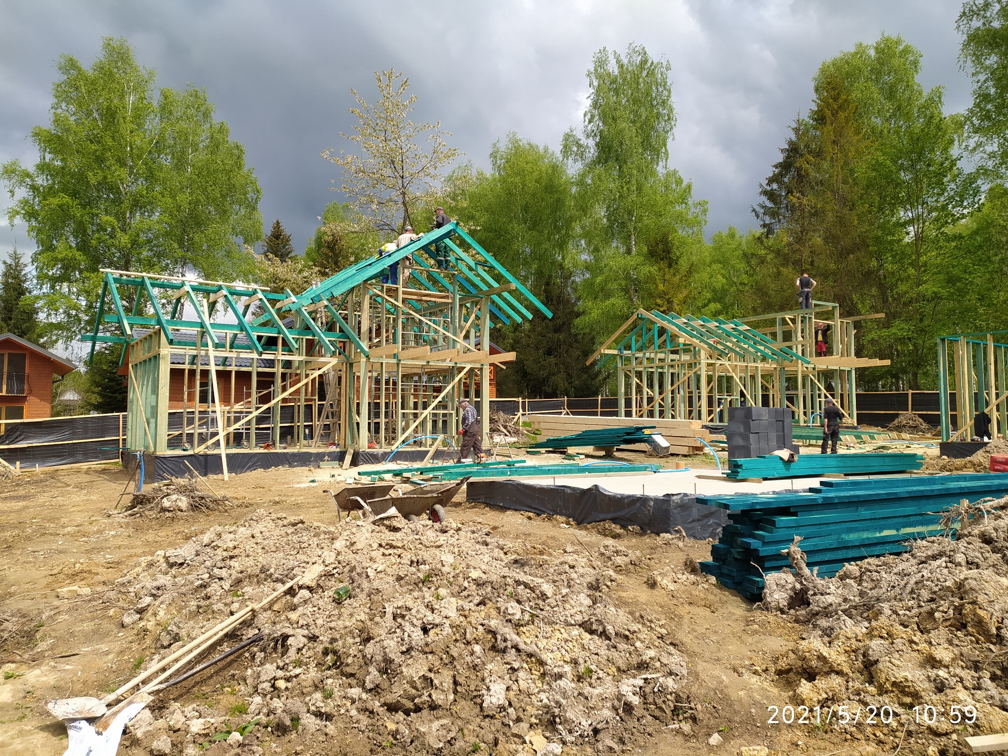 Wznoszenie dwóch domów szkieletowych w stanie surowym otwartym, z widocznymi drewnianymi konstrukcjami, pomalowanymi na zielono elementami dachu, pracownikami na rusztowaniach i stertą ziemi...