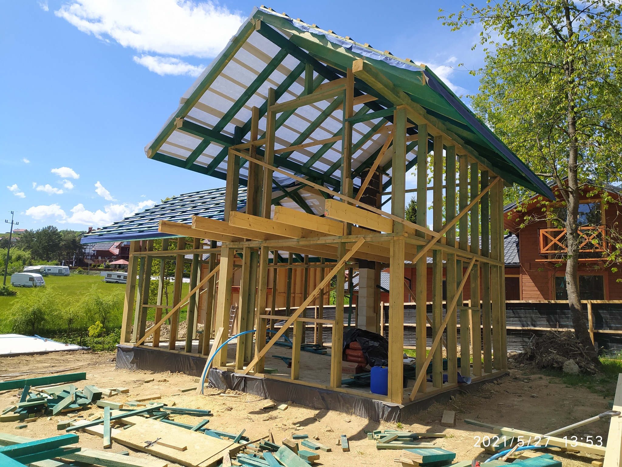 Budowa domu w konstrukcji szkieletowej: drewniany szkielet budynku z widocznymi elementami dachu i ścian, otoczony rozrzuconymi kawałkami drewna, na tle zielonej trawy i błękitnego nieba.