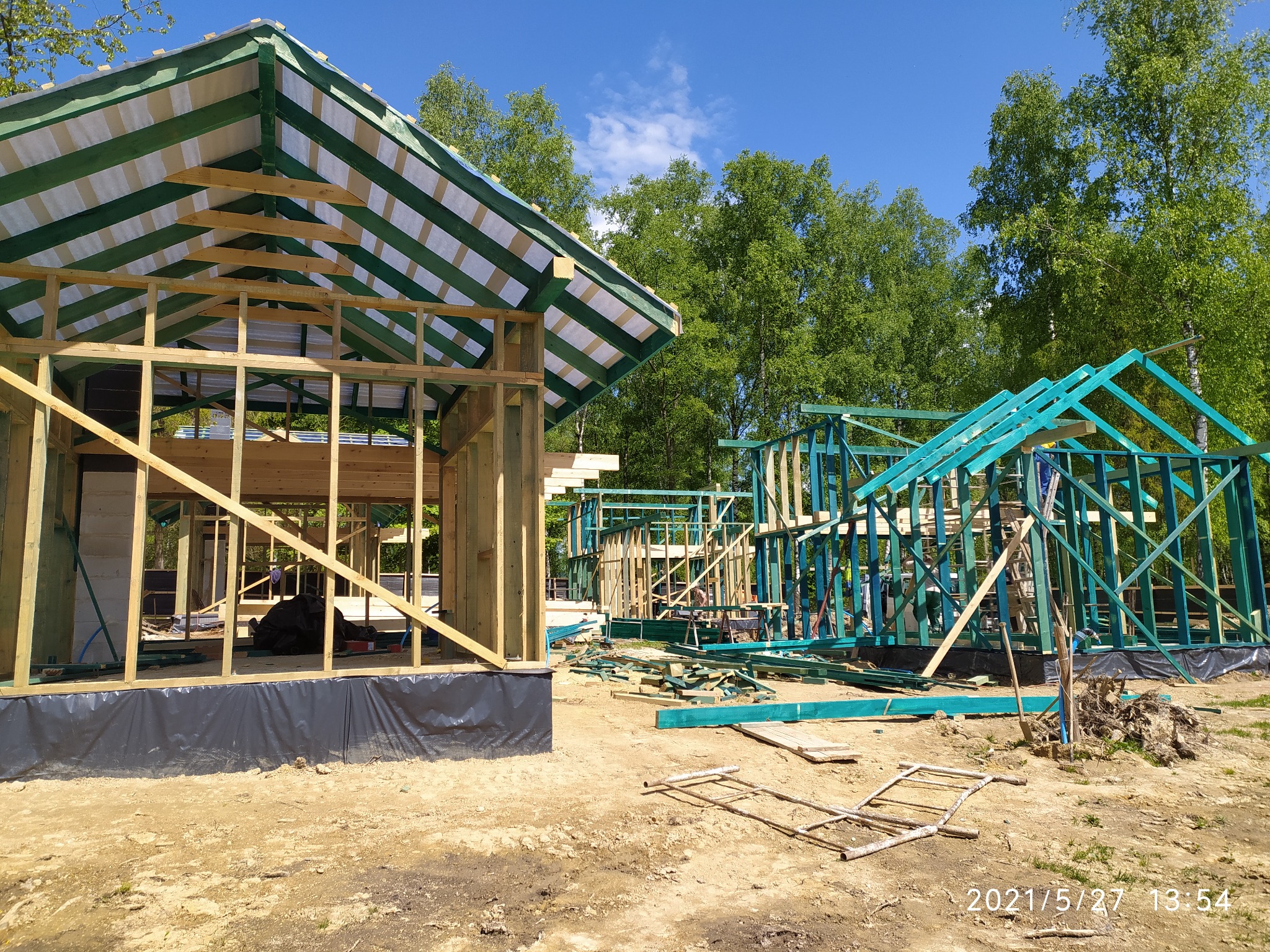 Wznoszenie drewnianej konstrukcji domu szkieletowego, widoczne elementy ścian i dachu w surowym stanie, na tle lasu i błękitnego nieba, z datą wykonania zdjęcia w prawym dolnym rogu.