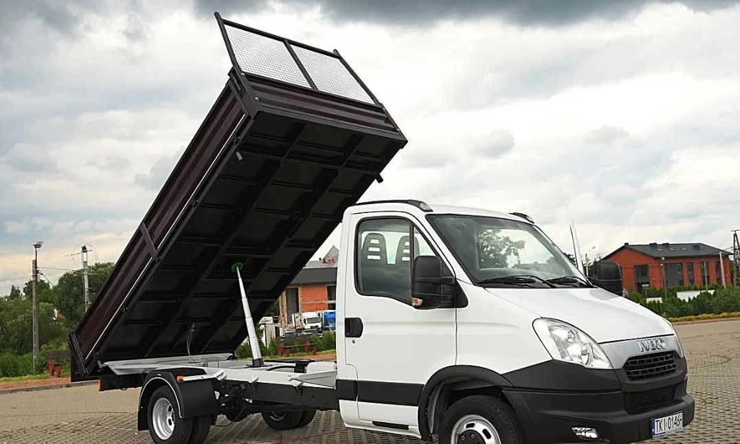 Biały samochód dostawczy z podniesioną, brązową skrzynią ładunkową, widok z boku na tle pochmurnego nieba.