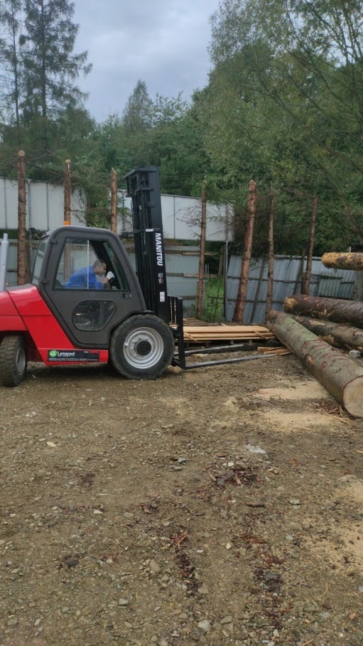 Czerwony wózek widłowy Manitou podnosi paletę z deskami na terenie składu drewna, obok leżą kłody drzew, w tle ogrodzenie i drzewa.