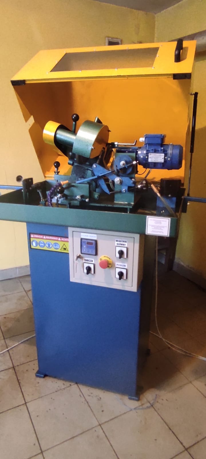 Stacjonarna szlifierka do ostrzenia narzędzi stolarskich z żółtą osłoną i niebieską podstawą, widoczne elementy sterujące i ostrzegawcze naklejki.