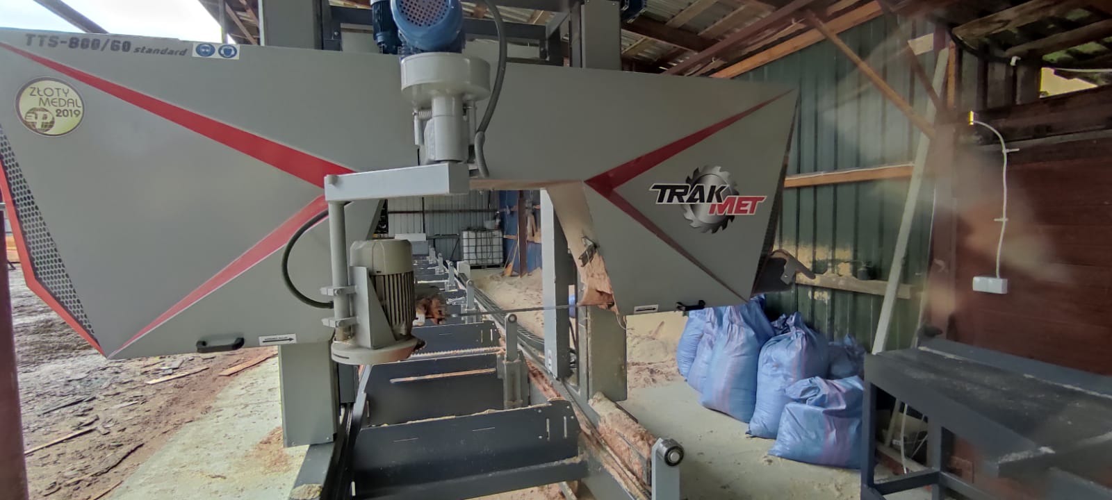 Profesjonalna piła taśmowa marki Trak-Met TTS-800/60 standard z nagrodą Złoty Medal 2019, ustawiona w hali z widocznymi workami z trocinami w tle, gotowa do obróbki drewna.