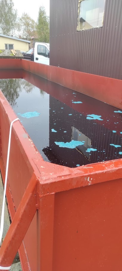 Czerwony, metalowy zbiornik wypełniony ciemną cieczą z niebieskimi plamami, prawdopodobnie impregnat do drewna, odbicie budynku w wodzie.