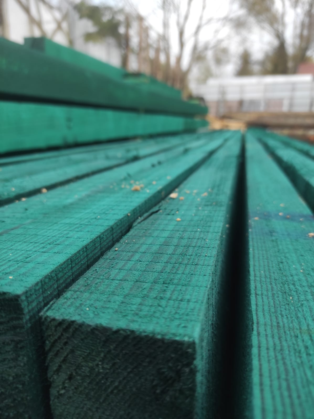 Stos impregnowanych na zielono kantówek z widoczną fakturą drewna i drobnymi wiórami, ułożonych równolegle w plenerze, z rozmytym tłem drzew i budynku.