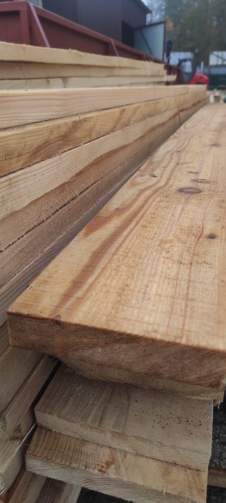 Stos świeżo przetartego, jasnego drewna budowlanego, widoczne słoje i niewielkie wióry na powierzchni, w tle zarys budynku gospodarczego i fragment maszyny budowlanej.