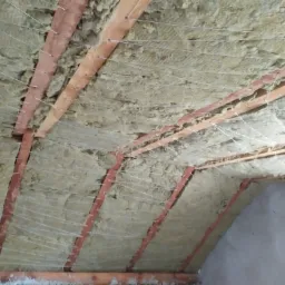 Izolacja poddasza z widocznymi belkami konstrukcyjnymi, wełną mineralną i siatką podtrzymującą, zabezpieczona sznurkiem przed dalszymi pracami wykończeniowymi.