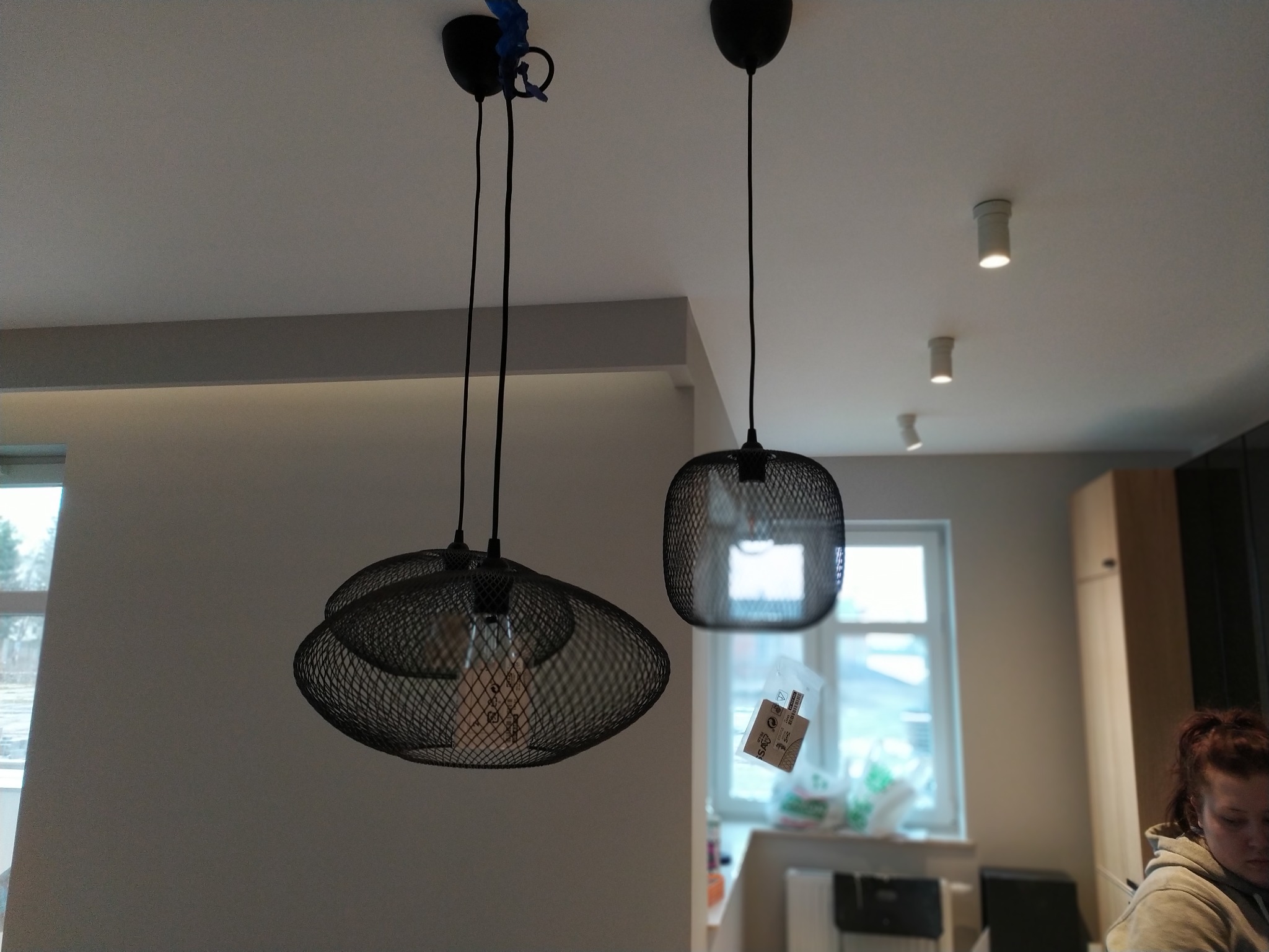 Dwa czarne metalowe abażury wiszące na przewodach, jeden w kształcie siatki, drugi bardziej pudełkowy, z widocznymi żarówkami Edisona w środku, montaż w trakcie, z niebieską taśmą zabezpieczającą.