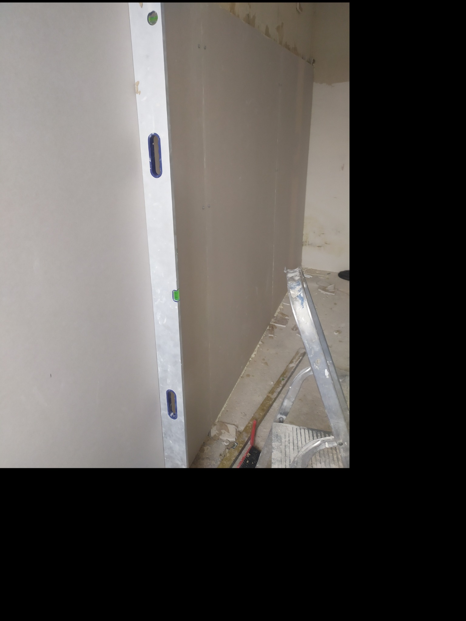 Ściana w trakcie wykańczania, widoczna zamontowana płyta z poziomnicą i fragment drabiny, ślady po remoncie na podłodze.