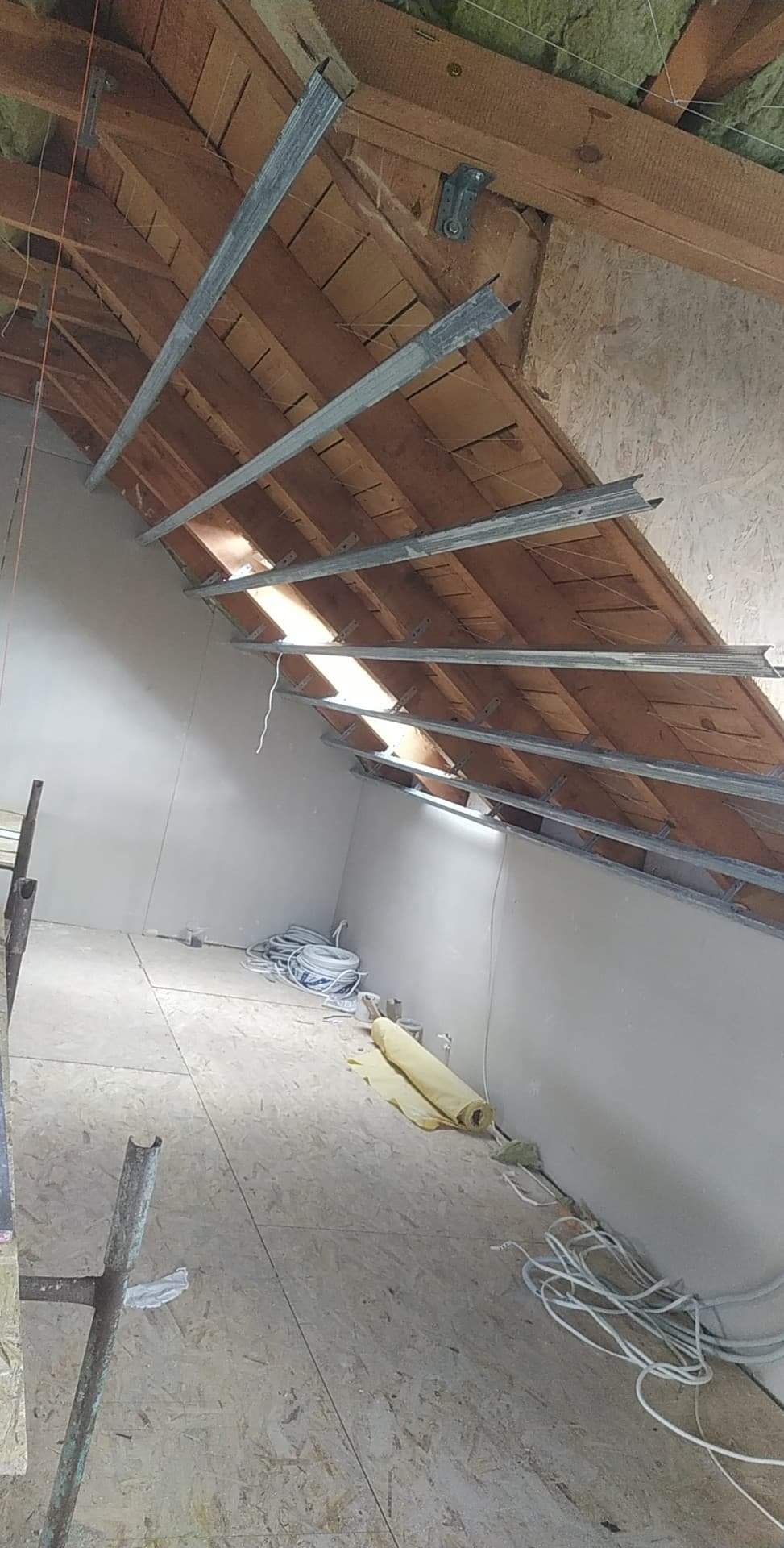 Montaż profili metalowych na drewnianej konstrukcji poddasza, widoczne przewody elektryczne i częściowo otynkowane ściany.