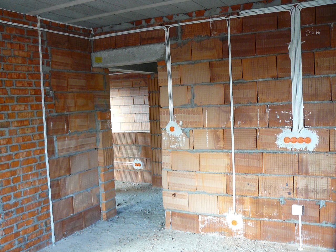 Wnętrze w trakcie prac: widoczne przewody elektryczne prowadzone po ścianach z cegły ceramicznej, puszki instalacyjne z pomarańczowymi zaślepkami, surowy stan wykończenia.