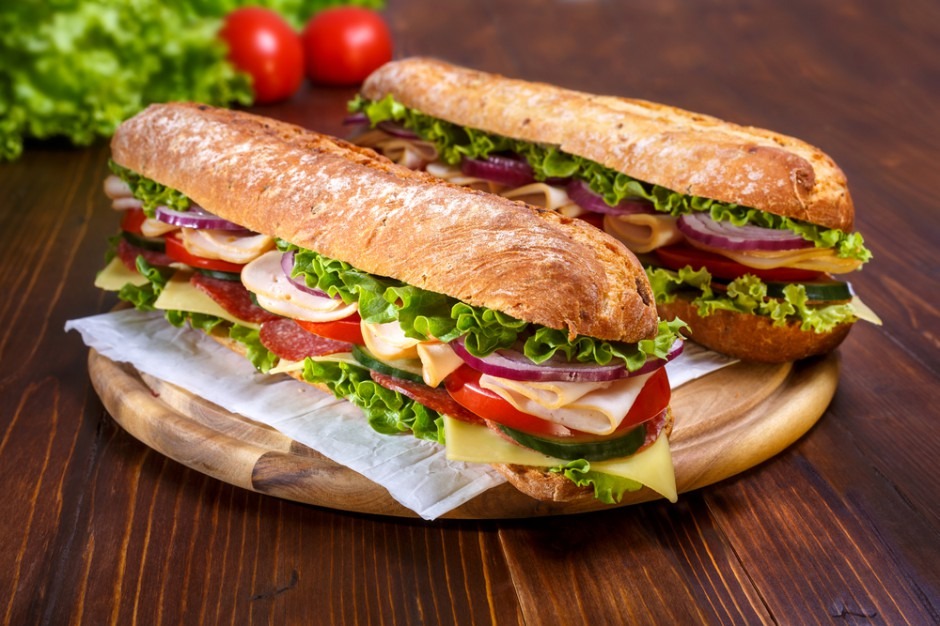 Dwa długie, przekrojone sandwicze na drewnianej desce, wypełnione serem, szynką, salami, pomidorem, ogórkiem, czerwoną cebulą i sałatą, z zieloną sałatą i pomidorami w tle.