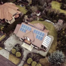 Widok z drona na dom jednorodzinny z panelami fotowoltaicznymi na dachu, otoczony zadbanym ogrodem i brukowanym podjazdem.