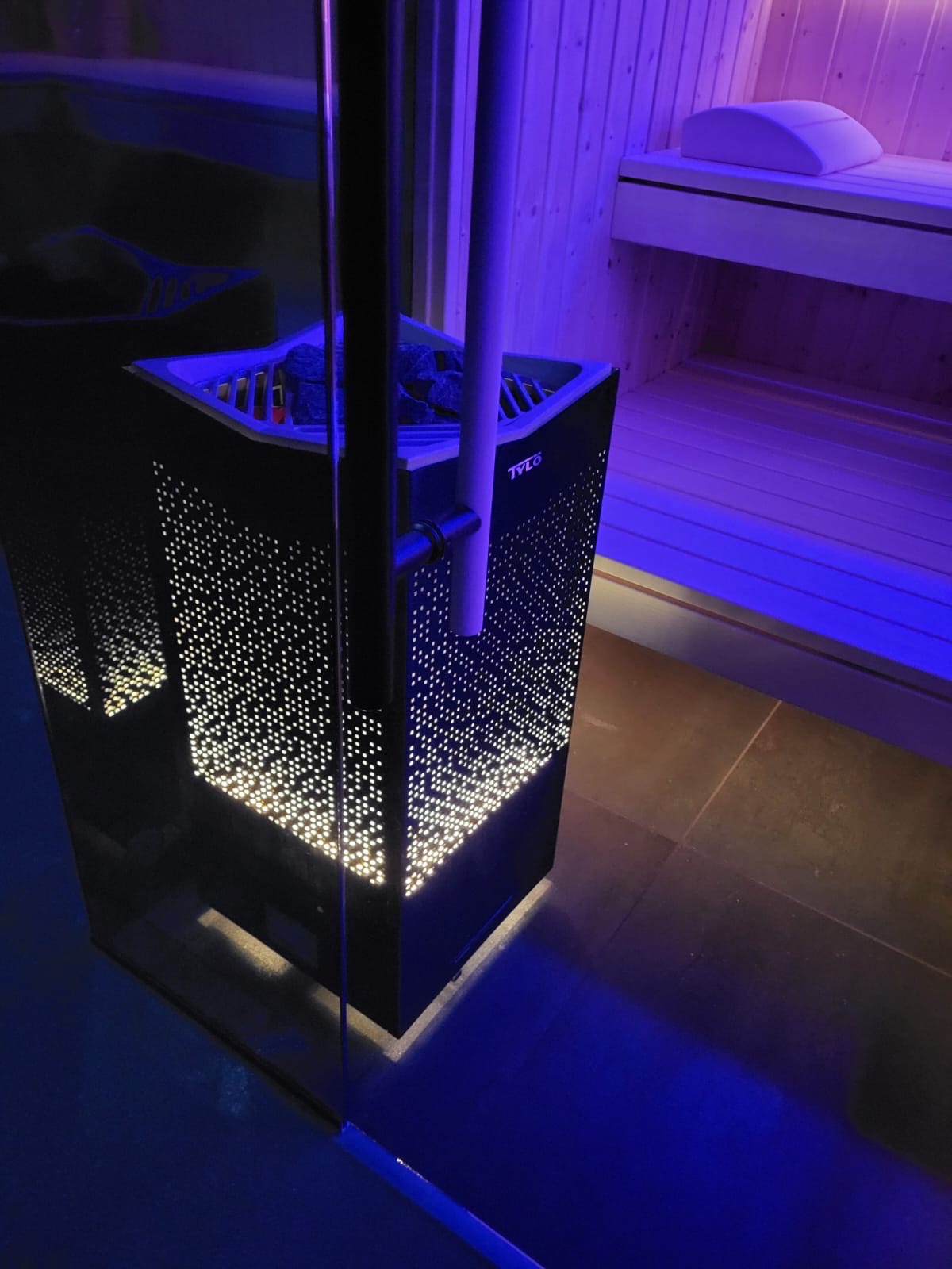 Elektryczny piec do ogrzewania kabiny w saunie, marki Tylo, z czarną, perforowaną obudową, podświetloną od wewnątrz, widoczne kamienie lawowe na górze, w tle drewniane ławki podświetlone niebieskim...