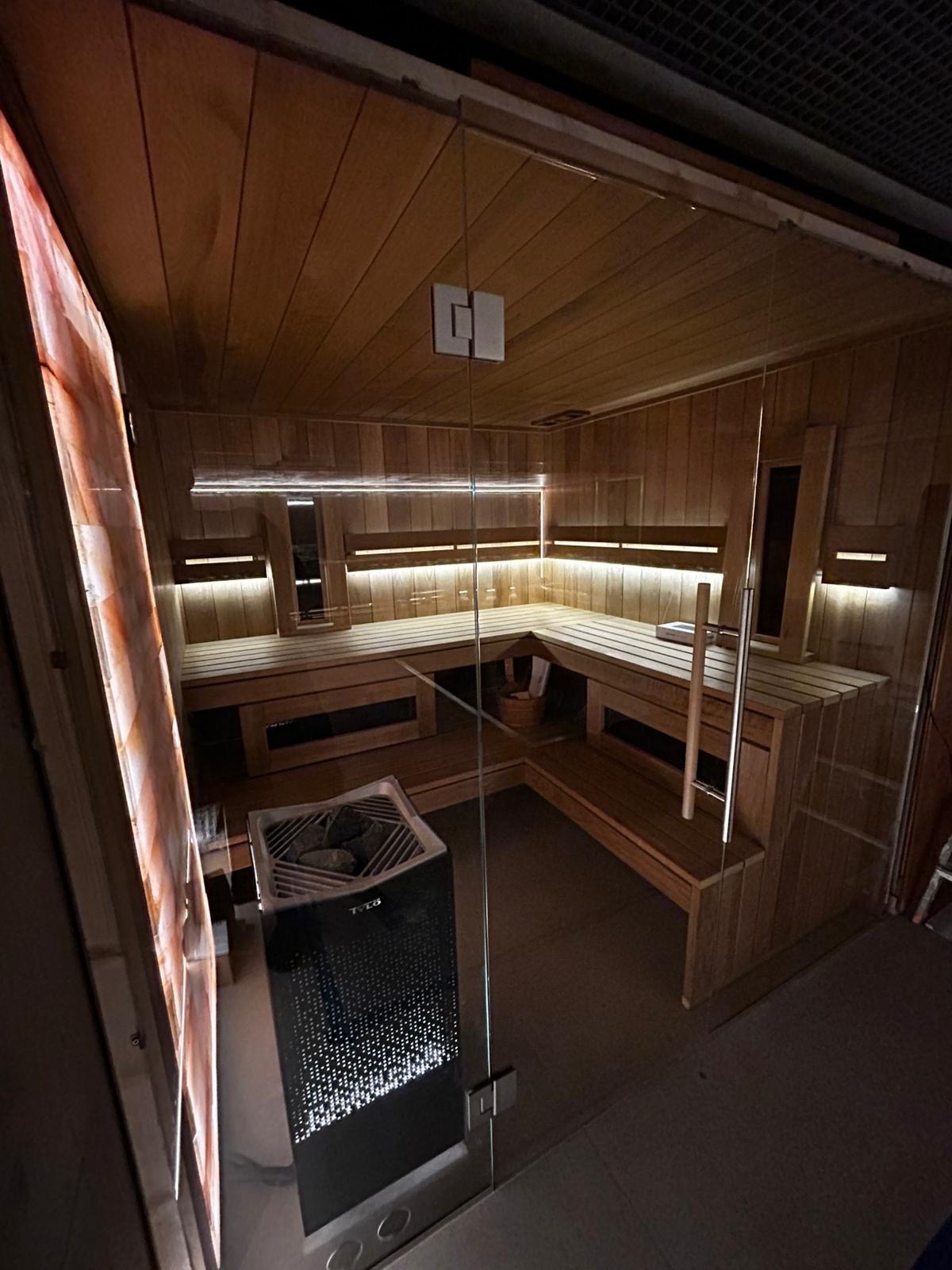 Wnętrze sauny z drewnianym wykończeniem, oświetleniem LED, szklanymi drzwiami i nowoczesnym piecem z kamieniami.
