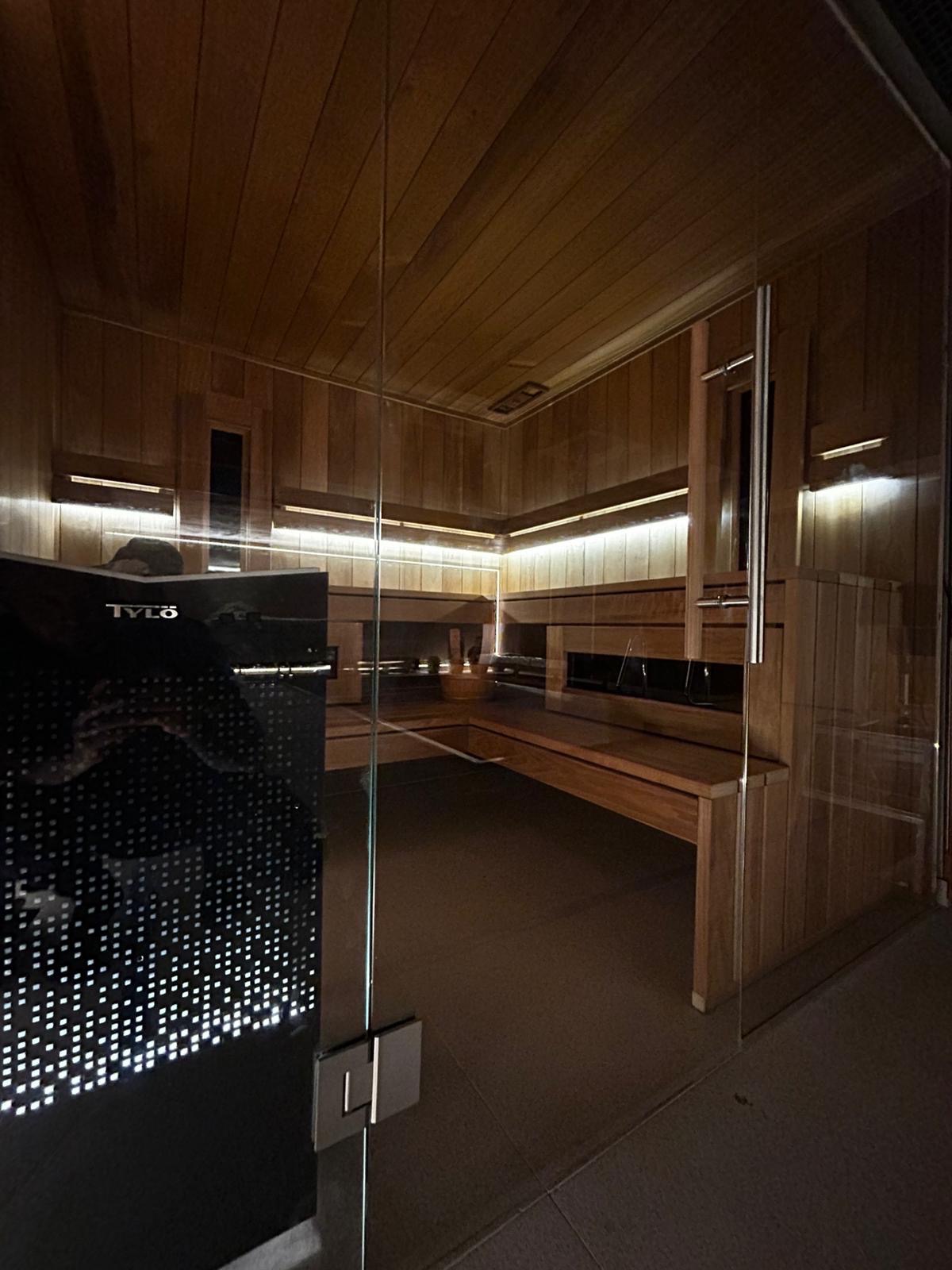 Wnętrze sauny z drewnianym wykończeniem, oświetleniem LED i przeszklonymi drzwiami z widocznym logotypem producenta na panelu sterowania.