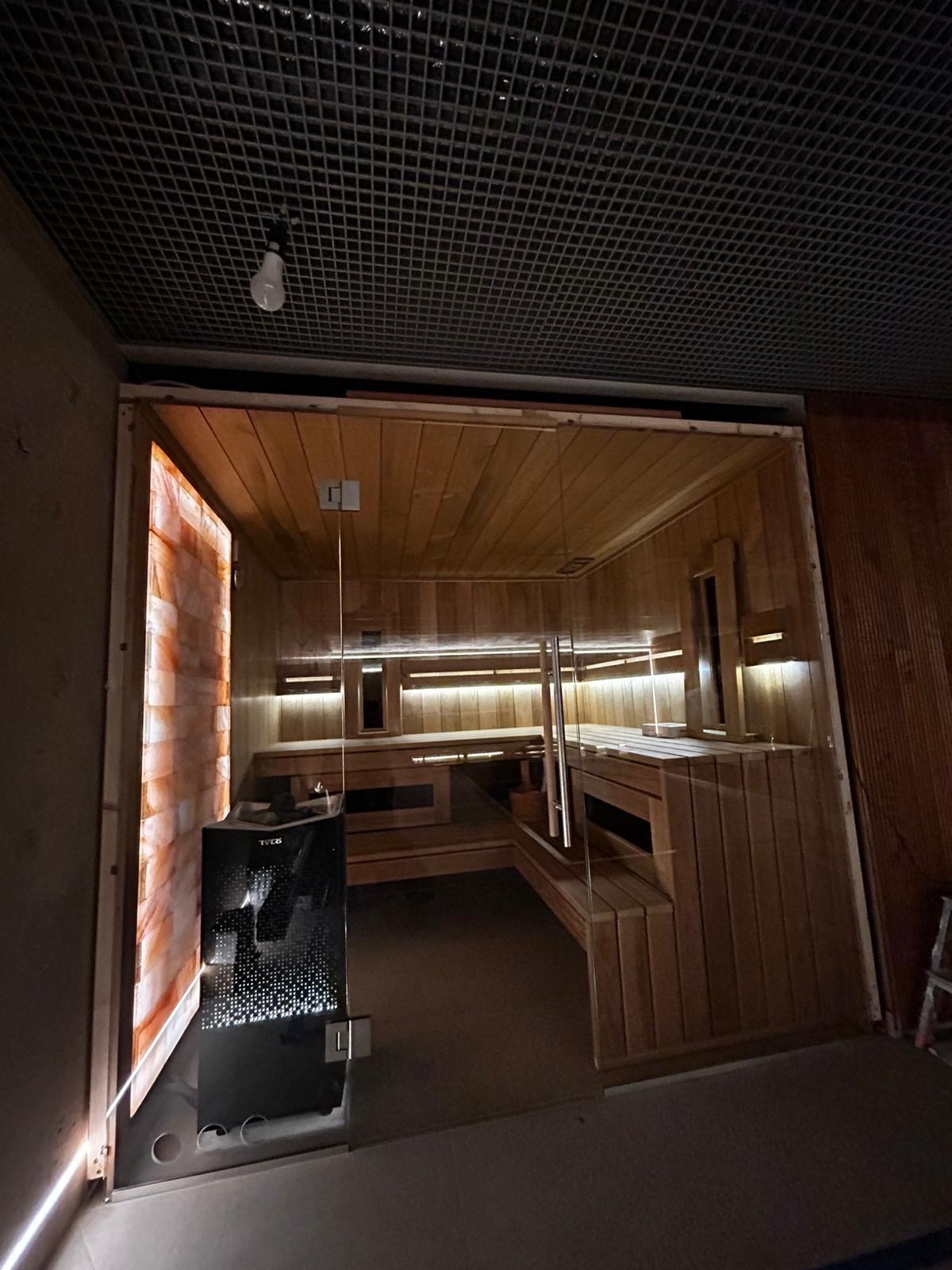 Nowoczesna sauna w Warszawie z drewnianym wykończeniem, przeszklonym wejściem i panelem z cegieł solnych po lewej stronie; widoczny piec do sauny marki Tylo.