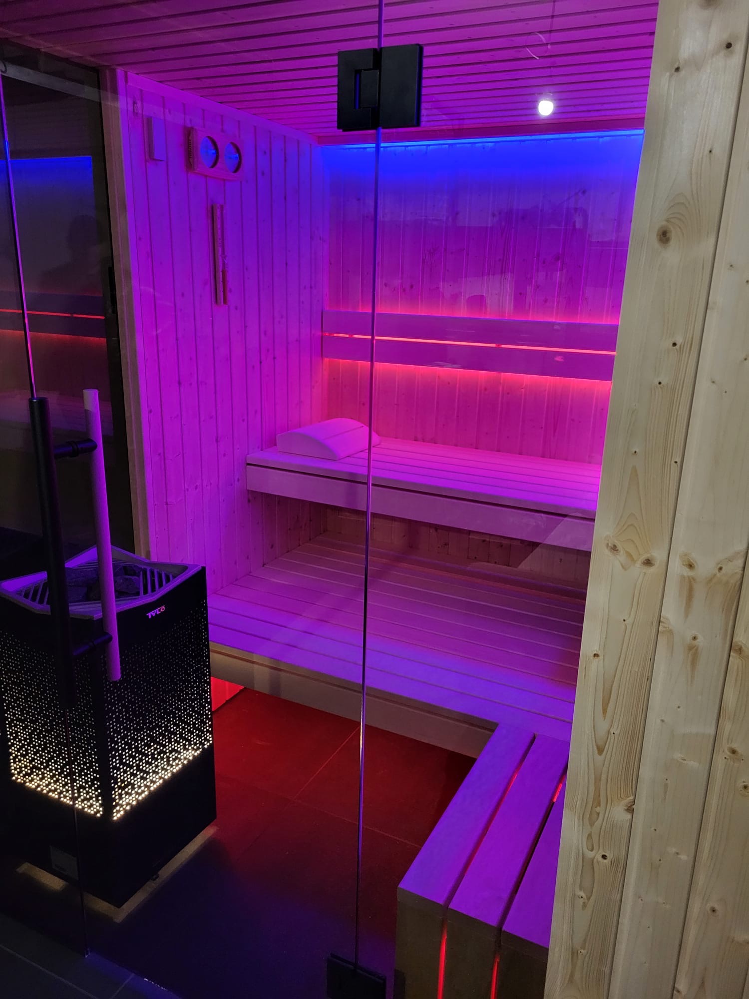 Wnętrze sauny z drewnianymi ławkami, oświetlone intensywnym fioletowym i czerwonym światłem, widoczny piec z ażurową obudową marki Tylo.