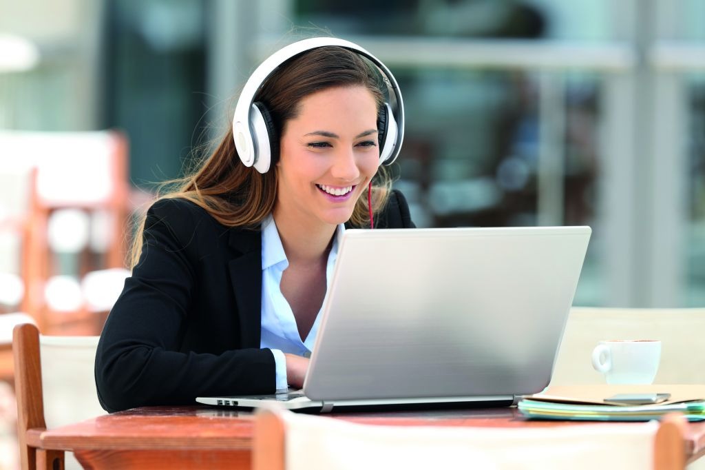 Uśmiechnięta kobieta w słuchawkach, w marynarce, korzystająca z laptopa przy drewnianym stole na zewnątrz; filiżanka kawy i dokumenty obok.