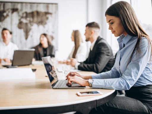 Kobieta w błękitnej koszuli pisze na laptopie podczas lekcji języka rosyjskiego, w tle mapa świata i inni uczniowie.