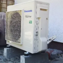 Biała jednostka zewnętrzna Panasonic Inverter R32 ustawiona na betonowych bloczkach przed białą ścianą z widocznym odpływem kondensatu.