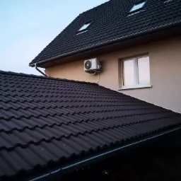 Zewnętrzna jednostka klimatyzacji zamontowana na ścianie domu z ciemną dachówką, widok z dołu pod kątem.