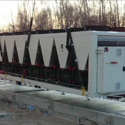 Montaż przemysłowego agregatu chłodniczego na zewnątrz budynku; widoczni pracownicy w kaskach i kamizelkach odblaskowych, betonowa podstawa i dźwig unoszący urządzenie.