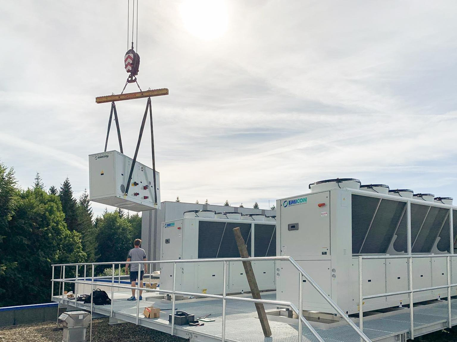 Montaż przemysłowego systemu klimatyzacji: dźwig unosi moduł klimatyzacyjny Emicon na platformie dachowej, w tle zainstalowane jednostki i pracownik nadzorujący.