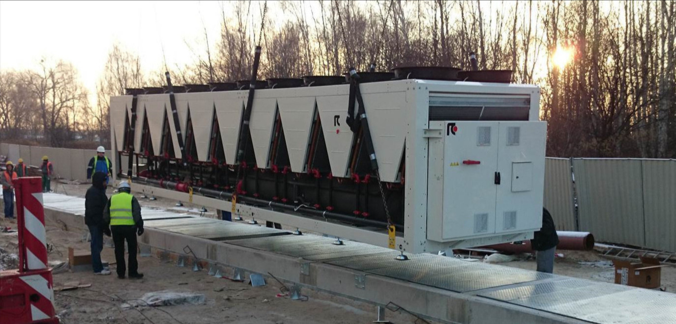 Montaż przemysłowego agregatu chłodniczego na zewnątrz budynku; widoczni pracownicy w kaskach i kamizelkach odblaskowych, betonowa podstawa i dźwig unoszący urządzenie.