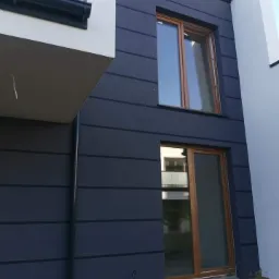 Fragment elewacji budynku z ciemnogranatową, poziomą fakturą, brązowe okna i białe elementy architektoniczne, widoczny fragment rynny.