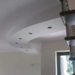 Wykończony sufit z wbudowanymi punktami świetlnymi i dekoracyjnym zaokrągleniem, widoczne schody spiralne i okno w tle.