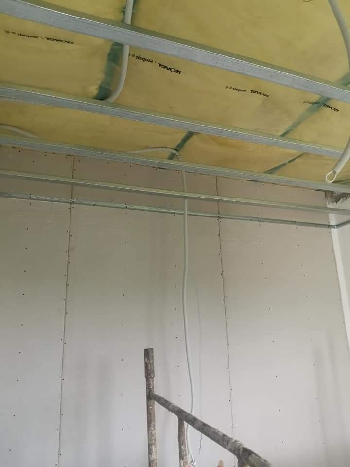 Montaż płyt gipsowo-kartonowych na ścianie i suficie z widoczną izolacją z wełny mineralnej oraz instalacją elektryczną, w tle fragment rusztowania.