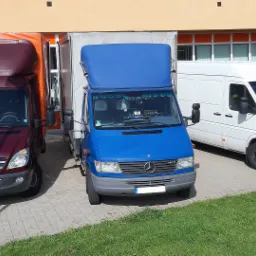 Trzy busy dostawcze Mercedes, zaparkowane obok budynku z pomarańczową fasadą, każdy w innym kolorze: bordowy, niebieski i biały, z widocznymi detalami jak lusterka i logo Mercedesa.